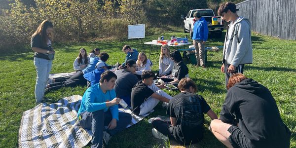 Group of people gathered outdoors on a sunny day, sitting on blankets and talking.