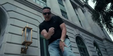 Man in sunglasses and black shirt posing energetically outside a stone building.