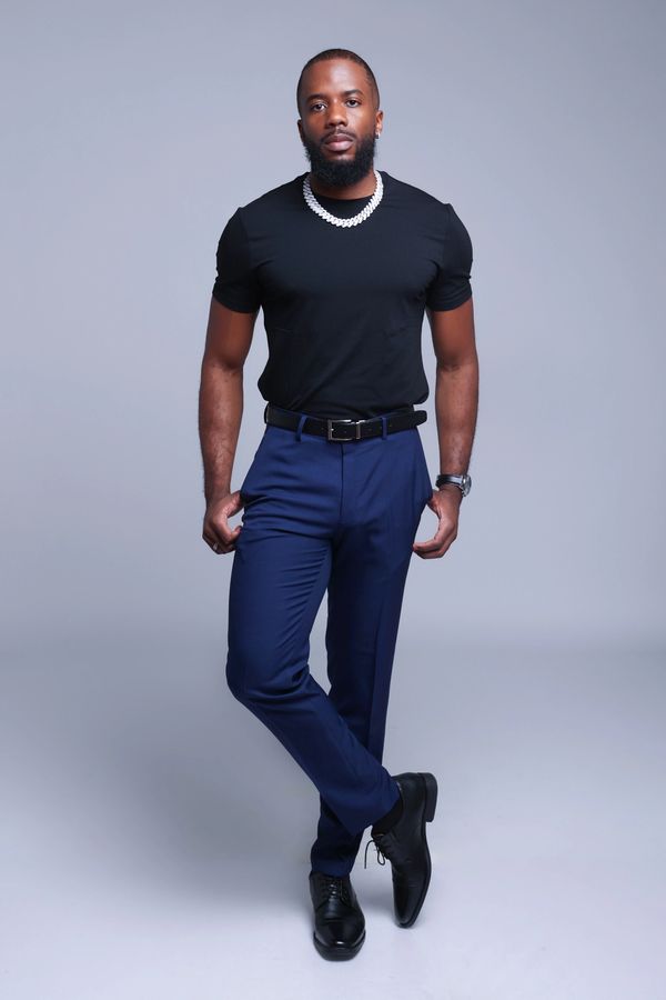 Man in black t-shirt and blue pants posing confidently against gray background.