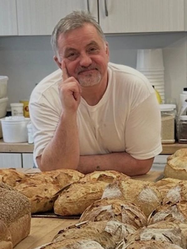 Man in white shirt poses with various freshly baked breads on table.
