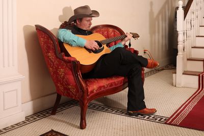 Man in cowboy hat playing guitar on a vintage red sofa.