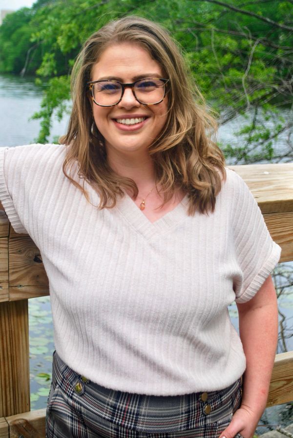 Smiling woman in glasses, standing outdoors by a wooden railing near water.