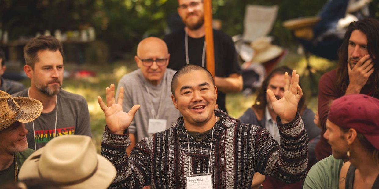 Man smiling and gesturing with hands in a group setting outdoors.