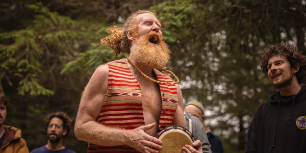 A joyful man with a red beard playing a drum outdoors.