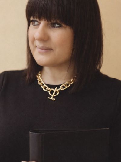 Woman in black holding a book, wearing a gold chain necklace, looking thoughtfully to the side.