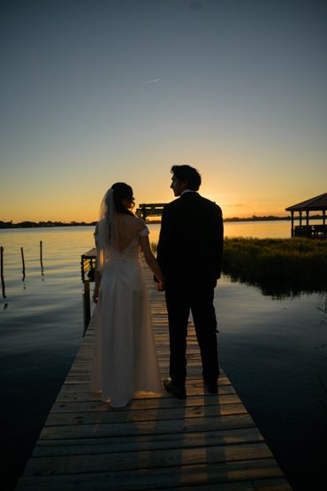 A couple in wedding attire holding hands on a dock at sunset.