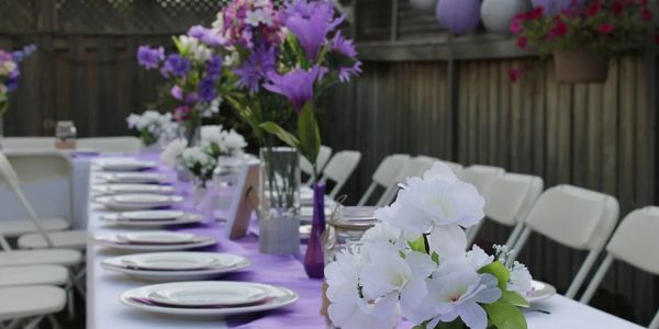Outdoor dining table decorated with purple and white flowers and a lavender tablecloth.
