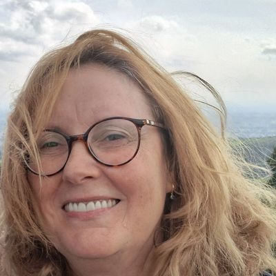 Smiling woman with glasses and windblown blonde hair outdoors under a cloudy sky.