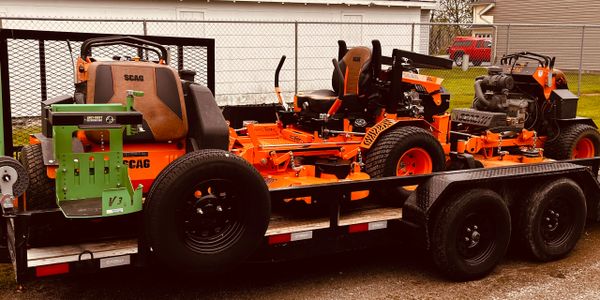 Orange SCAG riding lawn mowers loaded on a black trailer.