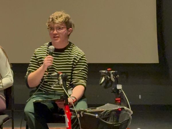 Mac speaks holding a microphone, sitting on a chair next to their walker