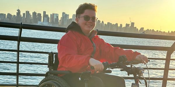 Mac sits smiling in their wheelchair near waterfront at sunset