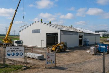 Construction site with workers installing roof panels on a large warehouse.