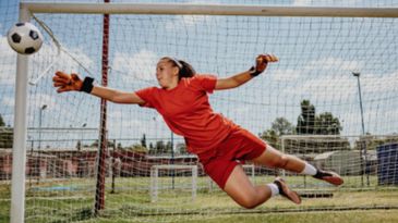 Female soccer goalkeeper diving to save a ball near the goalpost.