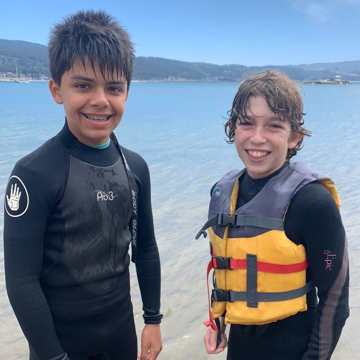 Two boys smiling in wetsuits by the water on a sunny day.