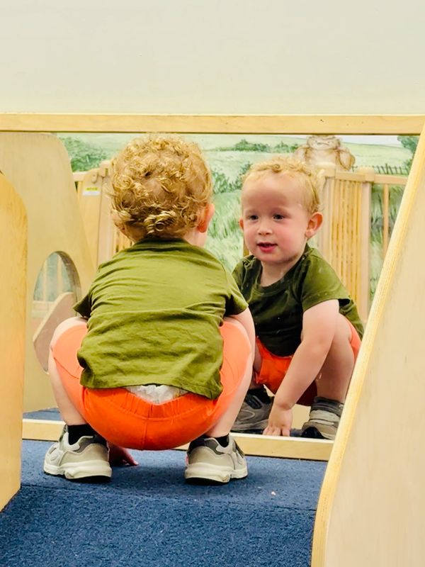 A toddler in orange shorts and green shirt looks at his reflection in a mirror.