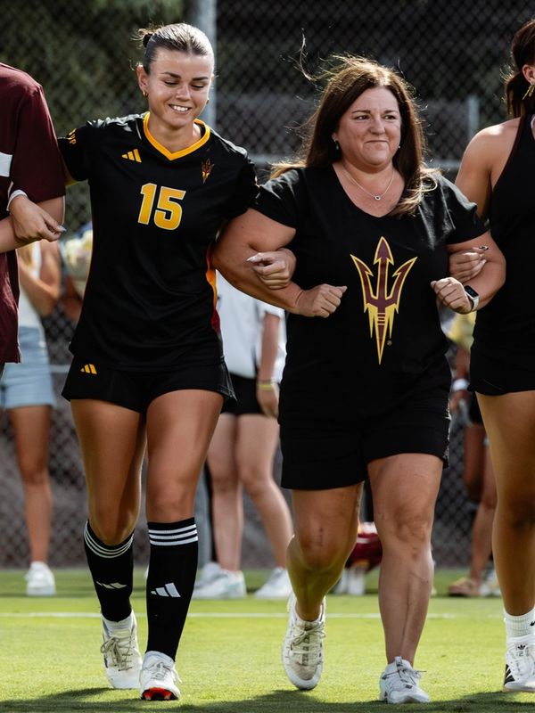 A female soccer player and a woman in team shirts linking arms on the field.