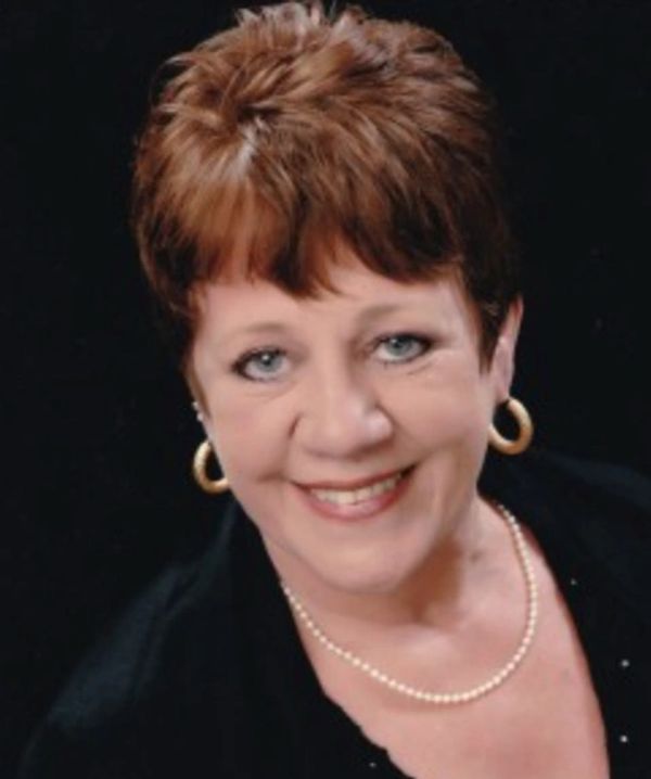 Smiling woman with short brown hair, pearl necklace, and gold hoop earrings against a black background.
