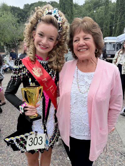 Young girl in Irish dance costume with trophy and sash, smiling next to an older woman.