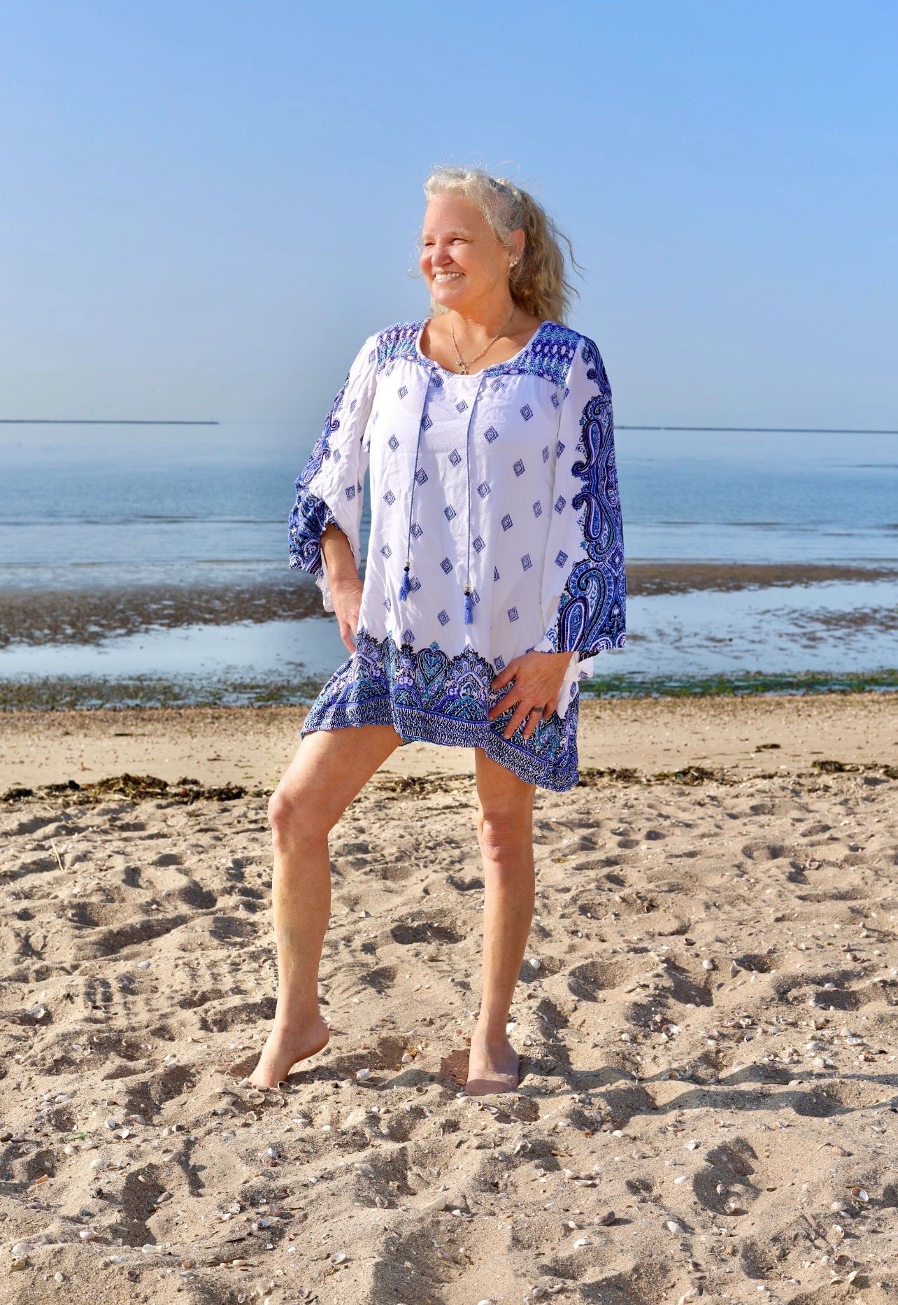 Smiling woman in a blue and white dress stands barefoot on a sandy beach by the sea.