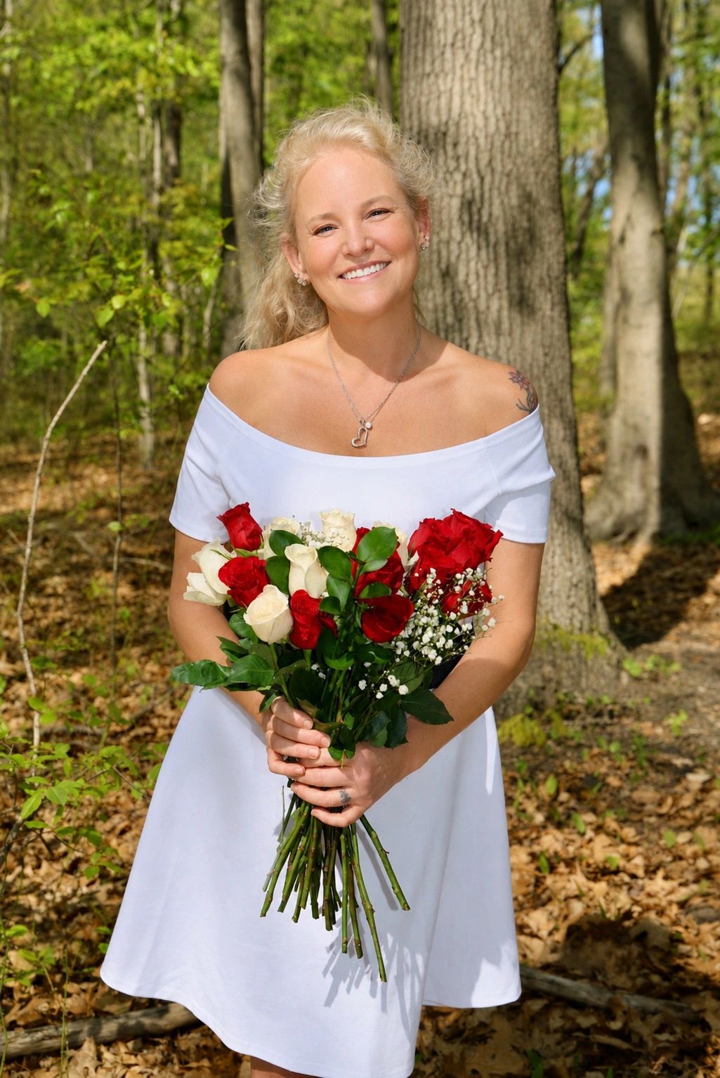 Smiling woman in white dress holding a bouquet of red and white roses in a forest.