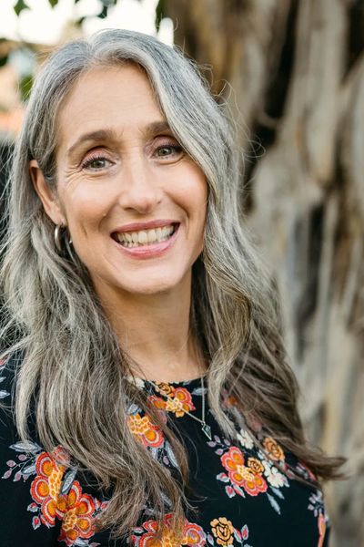 Smiling woman with gray hair in a floral top and jeans outdoors.