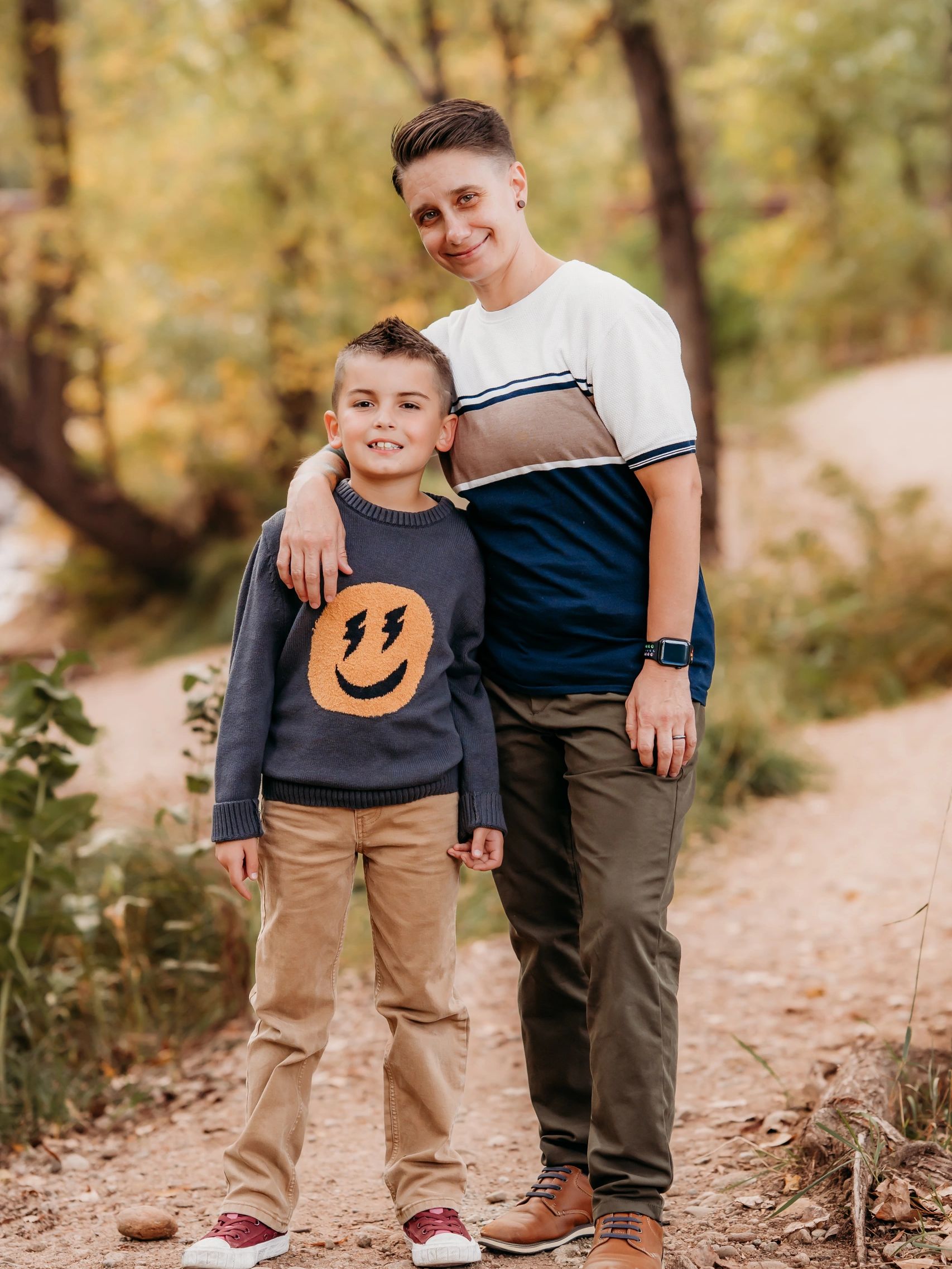 A smiling adult and child stand together outdoors on a dirt path in autumn.