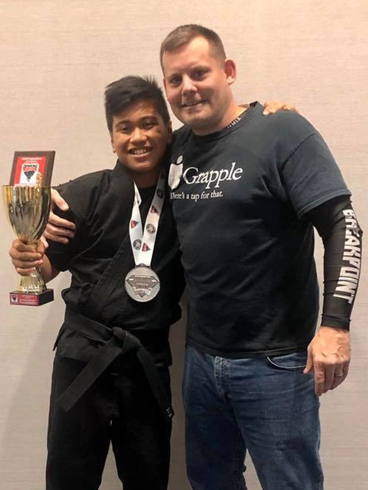 Two guys hugging. One guy wearing a black belt holding a trophy, plaque and a medal around his neck.