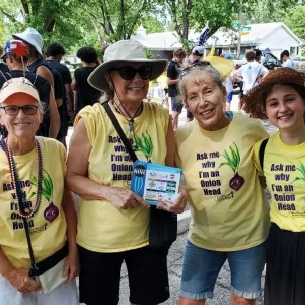 Four women wearing yellow shirts with 'Ask me why I'm an Onion Head' at an outdoor event.