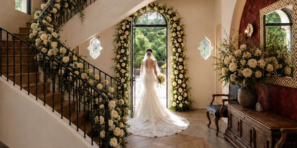 Bride in a lace gown stands at floral-decorated doorway in elegant venue.