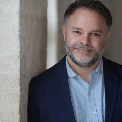 Middle-aged man with a beard smiles gently while leaning against a textured wall.