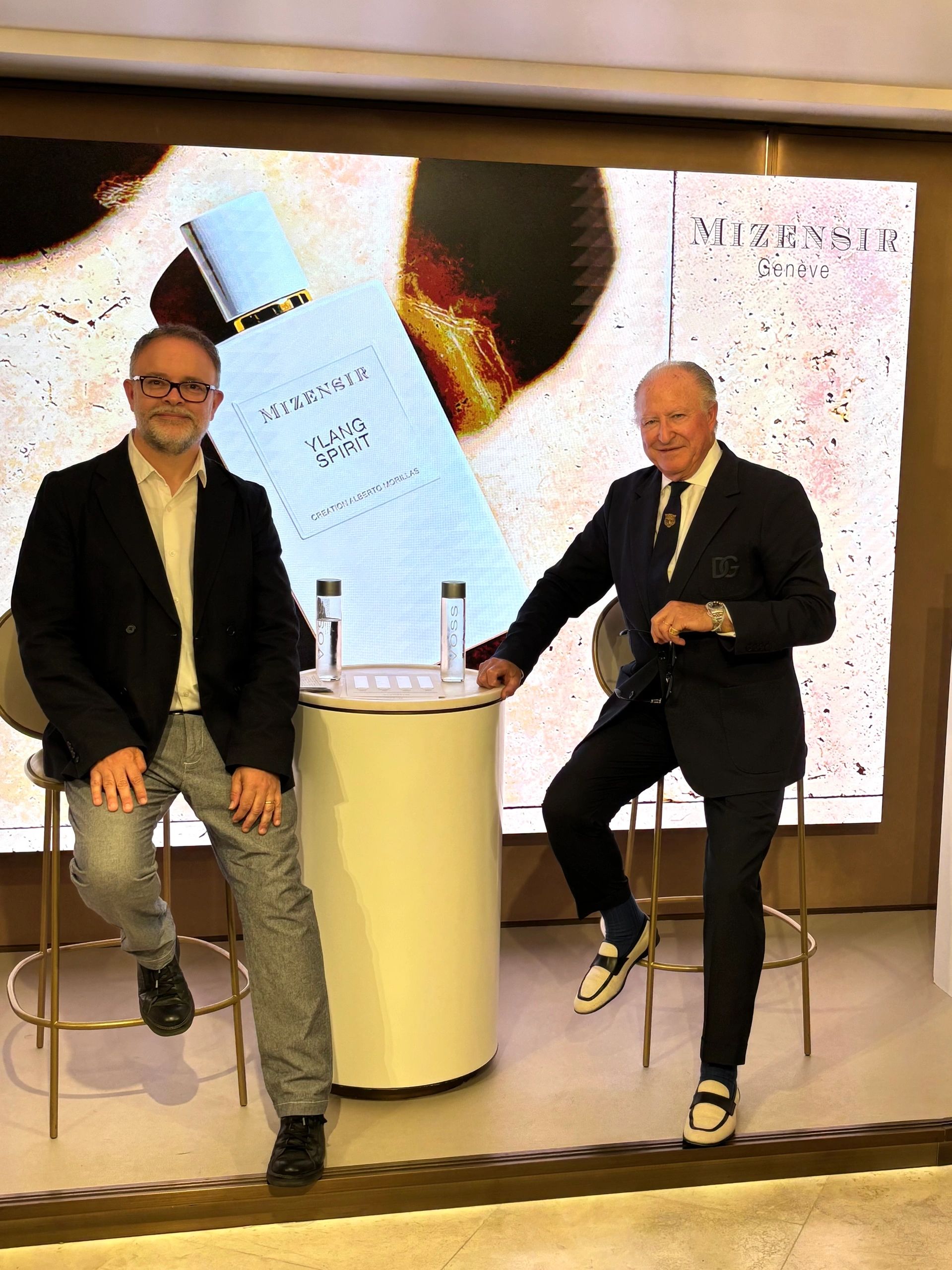 Two men sitting on stools in front of a Mizensir fragrance advertisement.