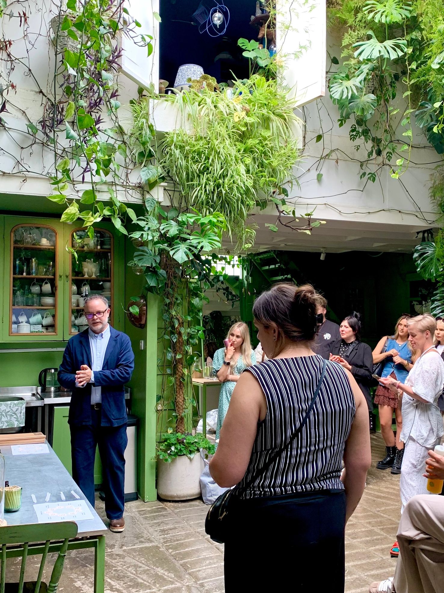 A man speaks to a group in a cozy, plant-filled room with green decor.