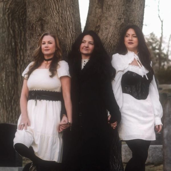 Three women in vintage-inspired outfits holding hands outdoors near a large tree.