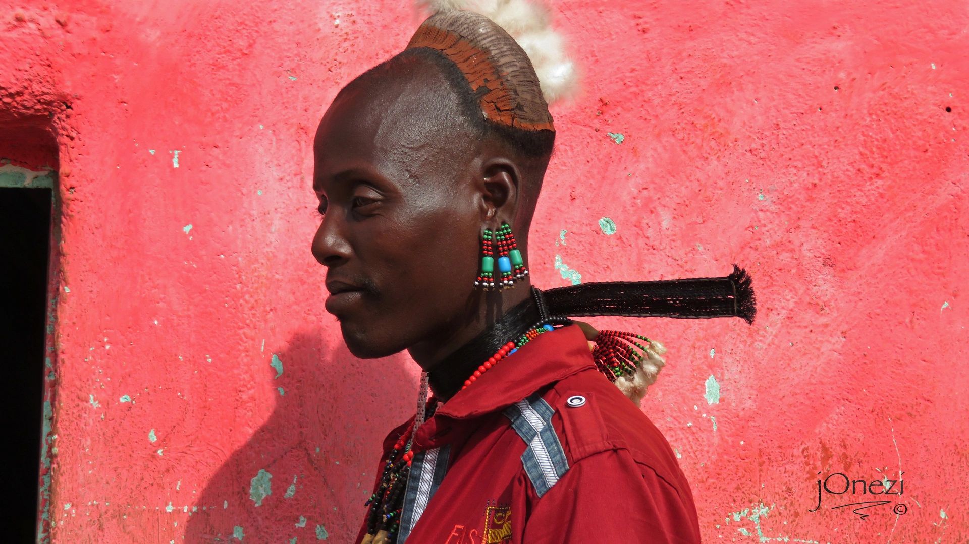 Profile of a man with traditional hairstyle and colorful beaded earrings against a red wall.