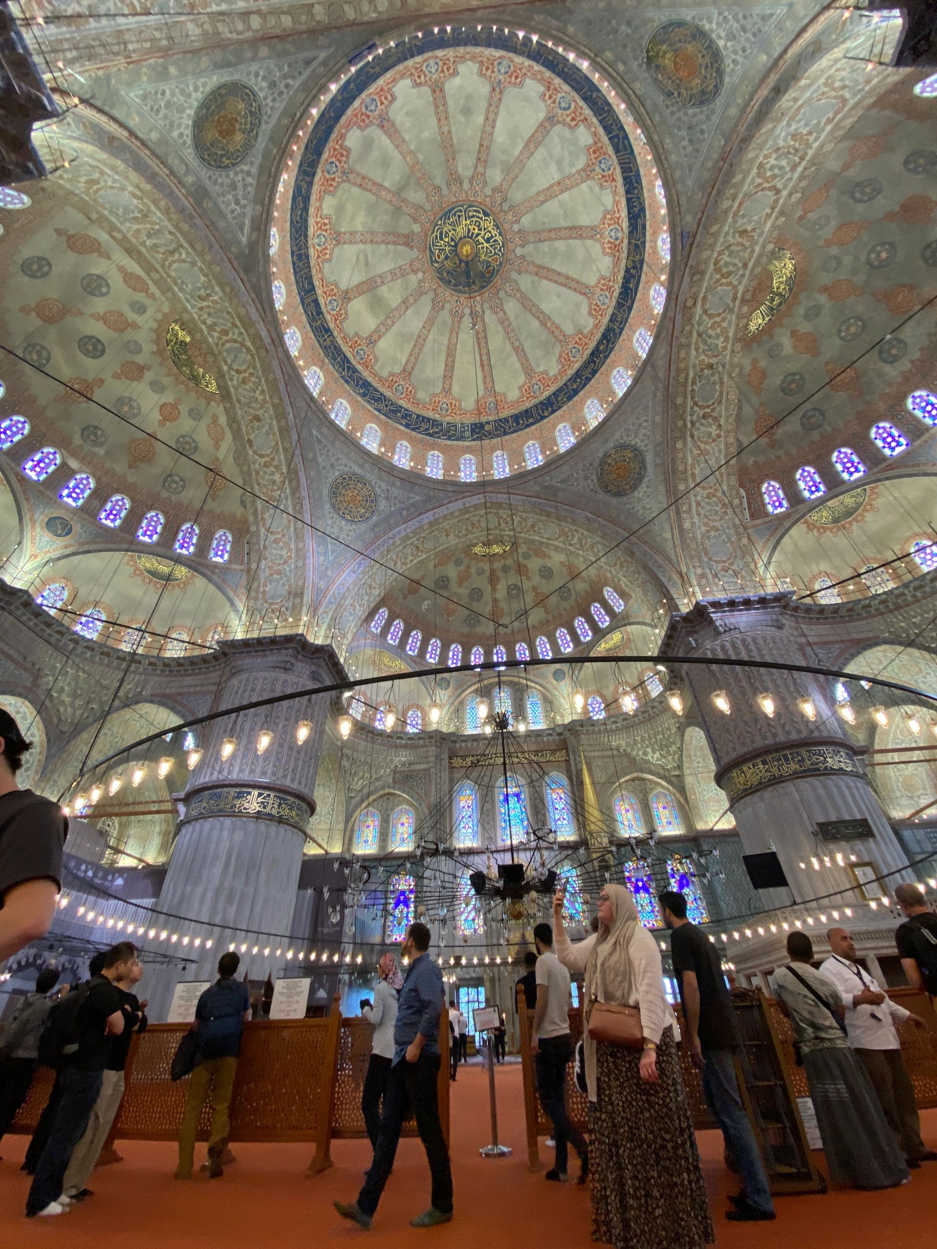 Interior of a grand mosque with intricate dome and visitors.