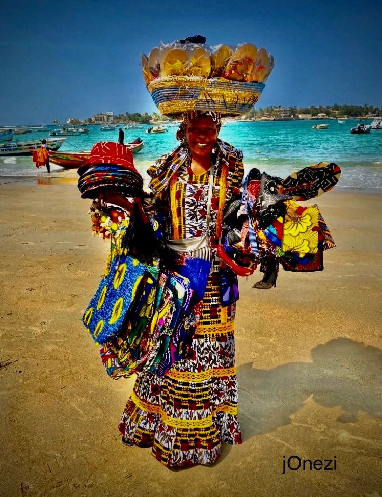 Woman in vibrant traditional attire selling colorful fabrics on a sunny beach.