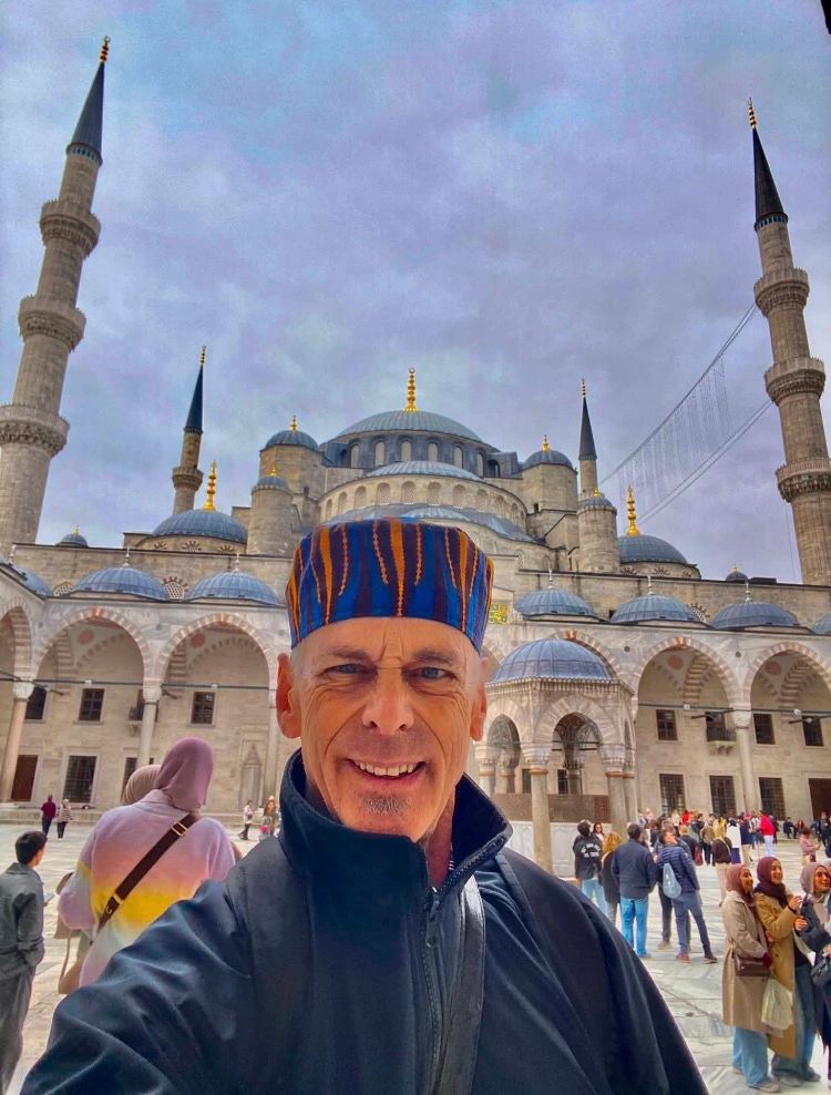 A man wearing a colorful hat takes a selfie in front of a mosque with minarets.