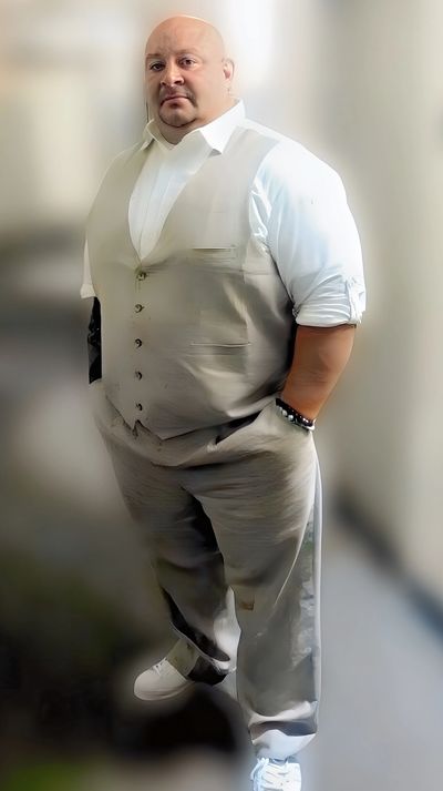 A man in a beige vest and pants with white sneakers stands confidently indoors.