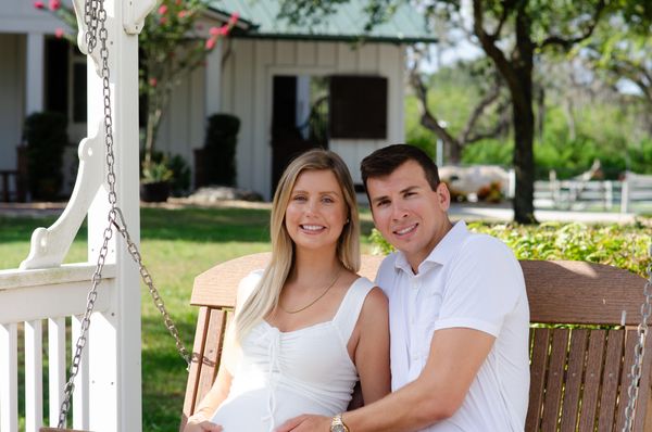 A pregnant woman sits smiling with her husband on a bench swing