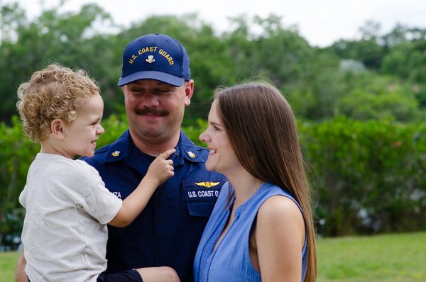 Family of 3 smiles at each other as the husband (wearing coast guard uniform) holds the young son