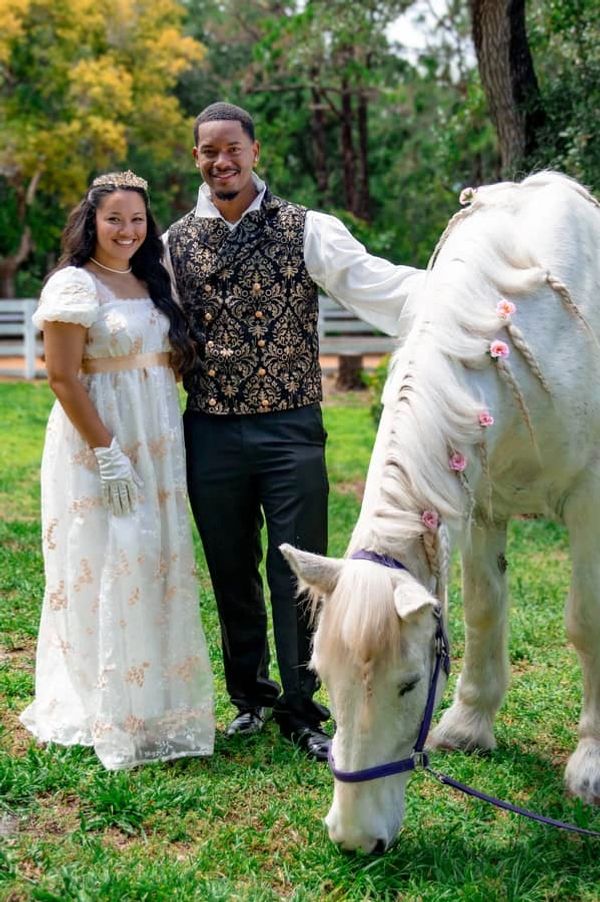 A couple in Regency era clothing stand with a white horse with pink flowers braided in its mane