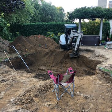 Excavation site in a backyard with a mini excavator and a pink folding chair.