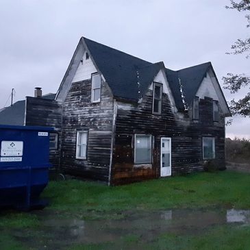Old weathered two-story house with peeling paint near a dumpster on a gloomy day.