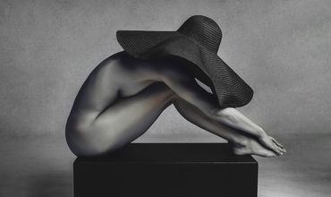 Artistic black and white photo of a nude figure wearing a large hat, sitting on a pedestal.