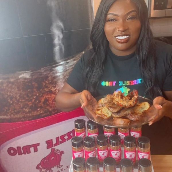 Smiling woman holds a plate of cooked food, surrounded by seasoning bottles.