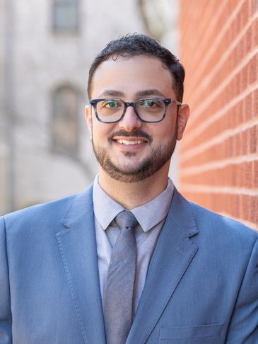 Man in glasses and blue suit smiling near brick wall.