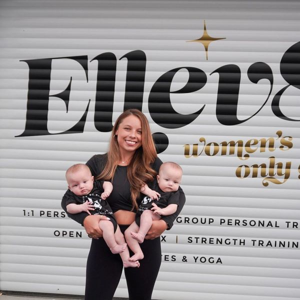 Smiling woman holding twin babies in front of a women's only gym sign.