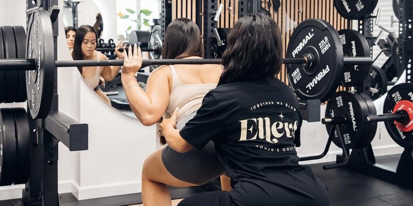 A woman squats with a barbell while another assists her in a gym.