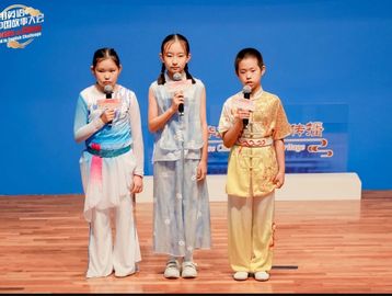 Three children in traditional attire sing on stage with microphones.