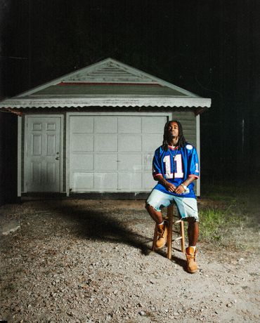 Man in sports jersey sitting on stool outside garage at night.